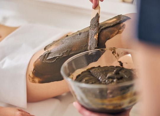 Close up of a wooden spatula and a glass bowl in hands of a cosmetologist. Body mask for the legs of young lady